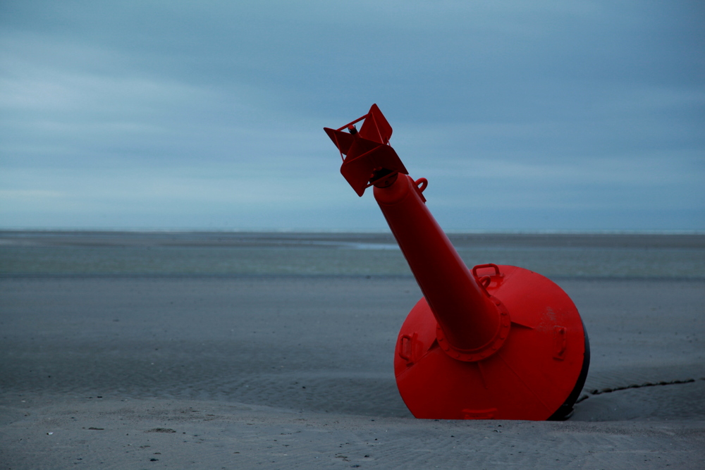 Posée sur le sable par marée basse, solidement arrimée au sol par une chaîne, la bouée de métal rouge indique le bord du chenal, utilisable par marée haute.

Le Touquet.